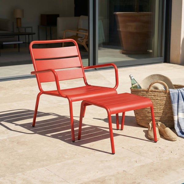 Roter Lounge-Sessel mit passender Fußablage aus Metall auf einer sonnigen Terrasse mit einem geflochtenen Korb und Schuhen daneben.