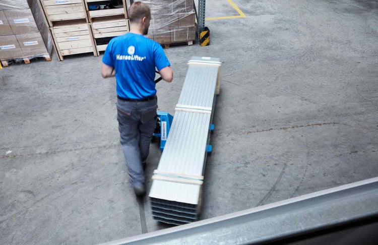 Mitarbeiter transportiert lange Metallprofile mit einem blauen Hubwagen in einem Lagergebäude.