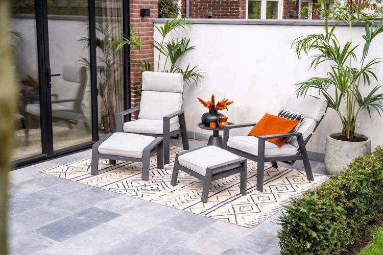 Terrassenset mit zwei Gartenstühlen mit hellen Polstern, zwei Fußhockern und einem runden Beistelltisch auf einem gemusterten Außenteppich im Garten.