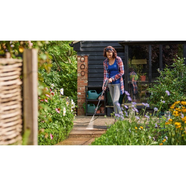 Frau reinigt ein blaues Rennrad mit einem kompakten grünen Hochdruckreiniger im Außenbereich vor einer dunklen Holzwand.