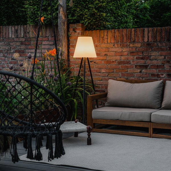 Moderne Außenstehleuchte mit Dreibeingestell und Schirm auf einer beleuchteten Terrasse mit Holzsofa und Pflanzen vor einer Backsteinmauer.