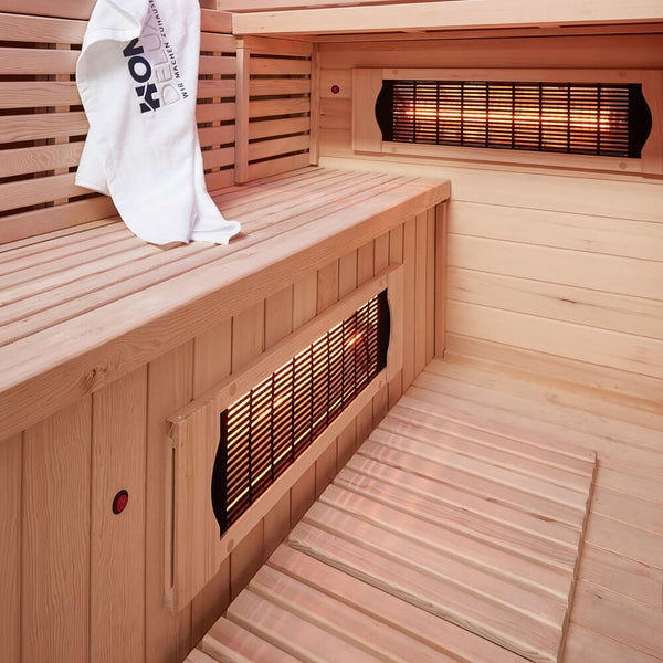 Innenansicht einer Infrarotkabine aus hellem Holz mit integrierten Strahlern, Sitzbank und einem Handtuch mit Logoaufdruck.