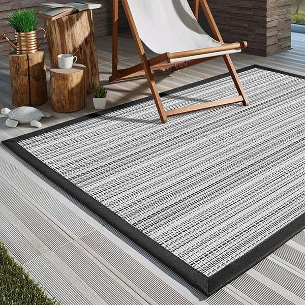 Rechteckiger Outdoor-Teppich mit gewebtem Muster und dunkler Bordüre auf einer Holzterrasse neben einem Liegestuhl aus Holz.