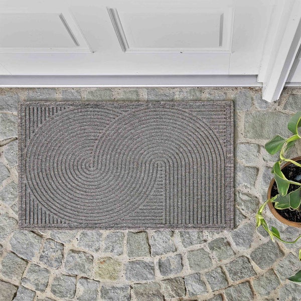 Graue Fußmatte mit Spiralmuster auf einem Steinboden vor einer Tür.