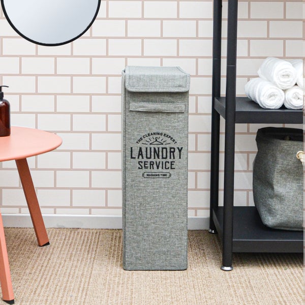 Schmaler Wäschekorb aus Stoff mit Aufdruck Laundry Service in einem hellen Badezimmer neben einem Regal mit Handtüchern.