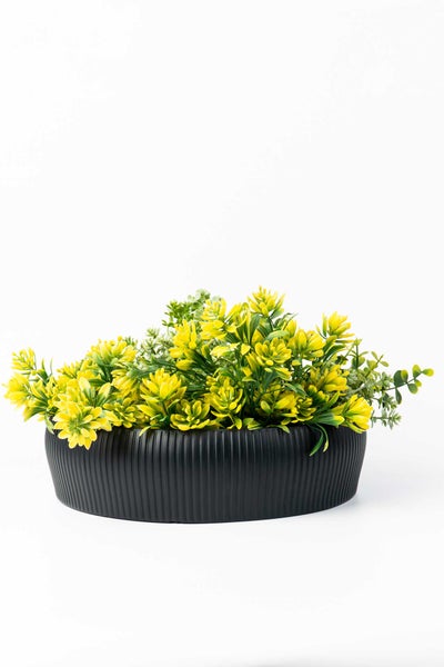 Künstliche Pflanze mit gelben Blüten in einer schwarzen, geriffelten Dekoschale.