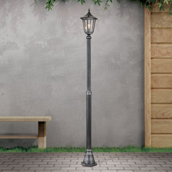 Klassische Außenstehleuchte aus Metall im Laternendesign auf einem gepflasterten Weg in einem Garten mit Holzbank und grauer Wand.