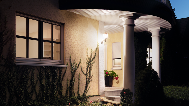 Beleuchtete Hausfassade mit klassischer Wandleuchte, Sprossenfenster und Efeu an der verputzten Wand neben einem Eingangsbereich mit Säulen.