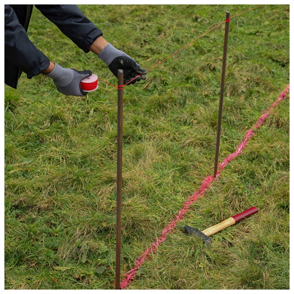Person spannt eine rote Markierungsschnur zwischen zwei Eisenstangen auf einer Rasenfläche. Ein Hammer liegt im Gras.