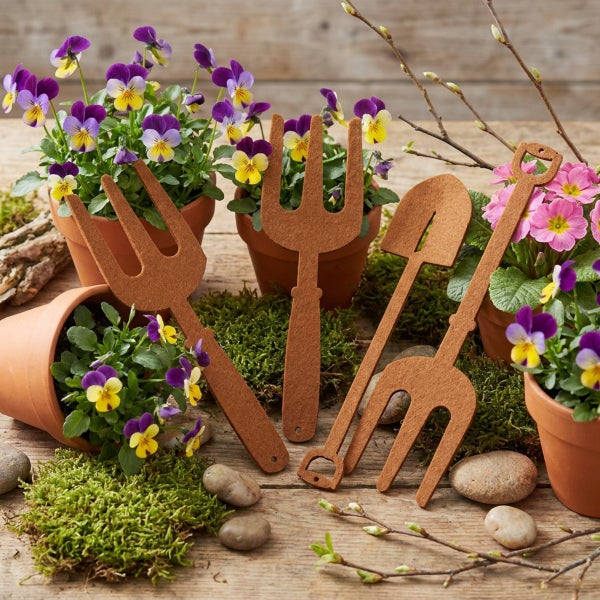 Deko-Gartengeräte aus Metall in Edelrost-Optik mit Blumen und Tontöpfen auf einem Holztisch als frühlingshafte Gartendekoration.