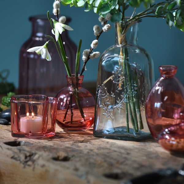 Verschiedene dekorative Glasvasen und ein Teelichthalter auf einer Holzplatte mit Frühlingsblumen und Zweigen dekoriert.