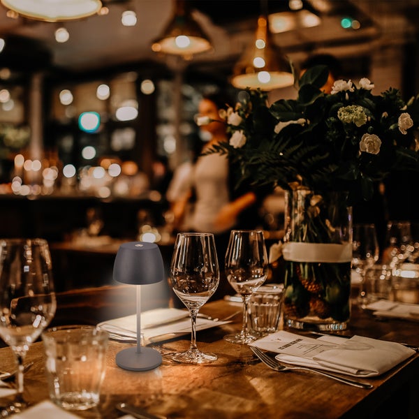 Moderne graue Akku-Tischleuchte auf einem gedeckten Restauranttisch mit Weingläsern und Blumen in warmer Atmosphäre.