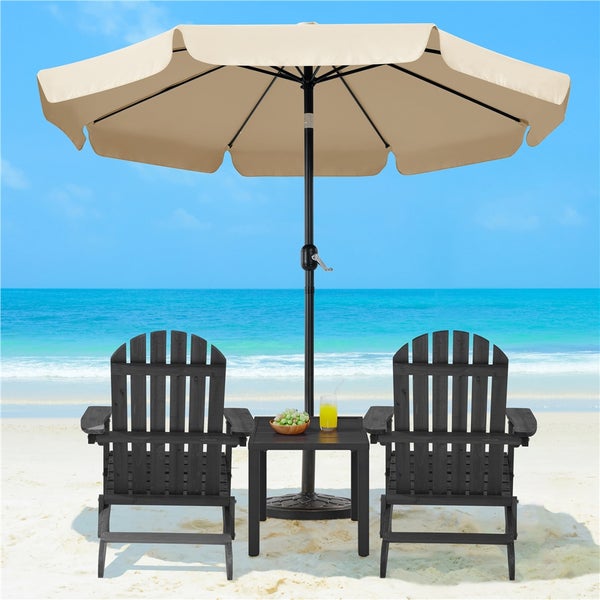 Zwei Adirondack-Stühle aus Holz, ein Beistelltisch und ein großer beiger Sonnenschirm auf einem weißen Sandstrand vor blauem Meer.