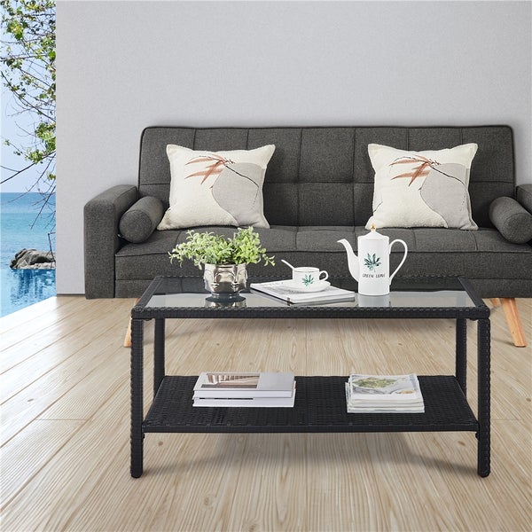 Rechteckiger Couchtisch aus schwarzem Polyrattan mit Glasplatte und Ablagefläche vor einem grauen Sofa in einem hellen, modernen Wohnzimmer.