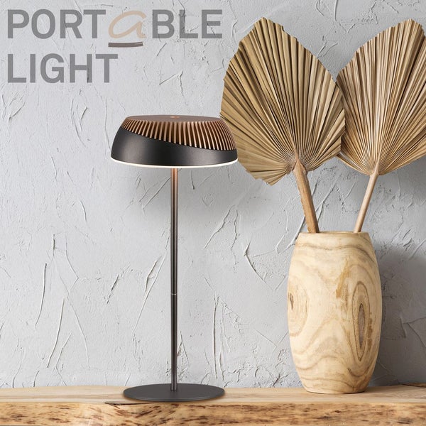 Tragbare Tischleuchte mit dunklem Metallschirm und Lamellen in Holzoptik auf einer Holzfläche neben einer Holzvase mit Trockenblumen.