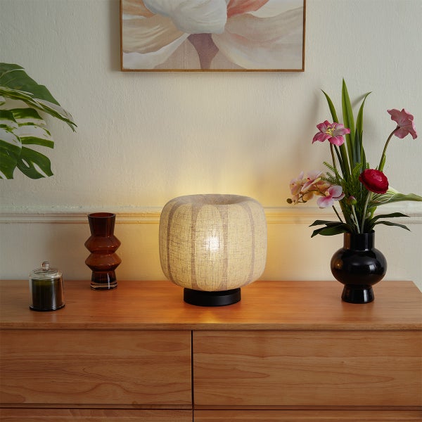 Tischleuchte mit rundem gestreiftem Stoffschirm auf einer Holzkommode neben Blumenvasen und Dekoration.