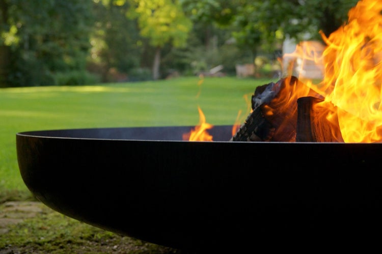 Eine dunkle Feuerschale aus Metall mit brennenden Holzscheiten und Flammen in einem Garten.