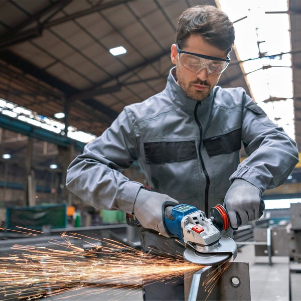 Ein Handwerker in grauer Arbeitskleidung und Schutzbrille bearbeitet ein Metallstück mit einem Winkelschleifer, wobei Funken fliegen.