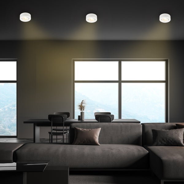 Drei weiße Einbaustrahler an der Decke in einem modernen Wohnzimmer mit Sofa, Esstisch und großen Fenstern mit Bergblick.