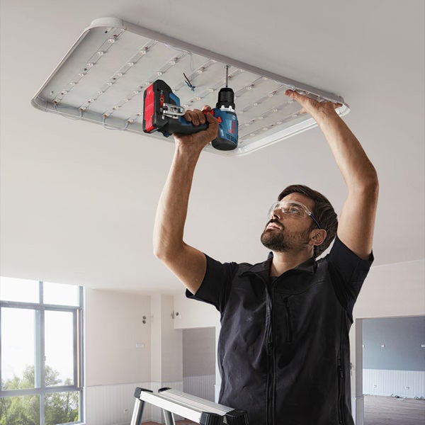 Handwerker mit Schutzbrille montiert eine LED Deckenleuchte mit einem Bosch Akkuschrauber an der Zimmerdecke