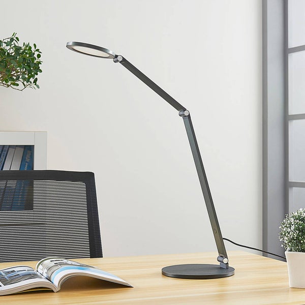 Verstellbare LED Schreibtischlampe mit rundem Leuchtkopf auf einem Holztisch in einem hellen, aufgeräumten Arbeitszimmer.