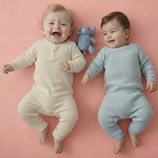 Zwei lächelnde Babys in cremefarbenen und hellblauen Strickstramplern liegen auf einer strukturierten Matte neben einem kleinen Stoffelefanten.