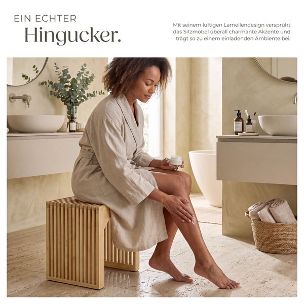 Eine Frau im Bademantel sitzt auf einem Holzhocker mit Lamellendesign in einem hellen, modern eingerichteten Badezimmer.