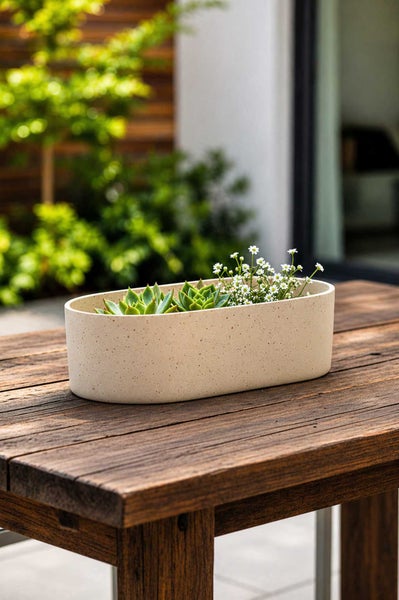 Ovaler Pflanzkasten mit gesprenkelter Oberfläche auf einem Holztisch, bepflanzt mit Sukkulenten und kleinen Blumen im Außenbereich.