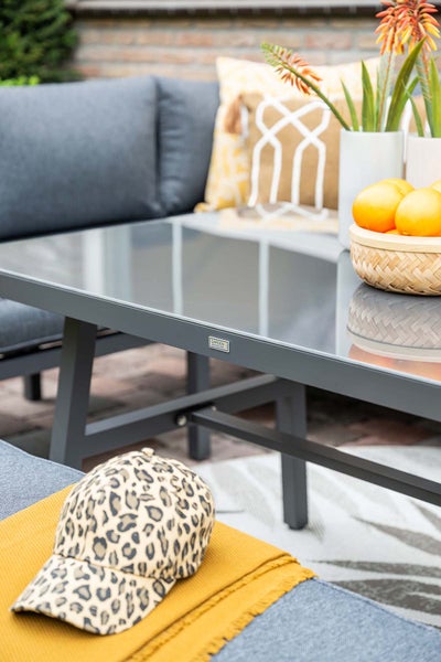 Loungetisch mit Glasplatte und Metallgestell in einem hellen Außenbereich mit Sofa und Dekoration sowie Garden Impressions Logo.