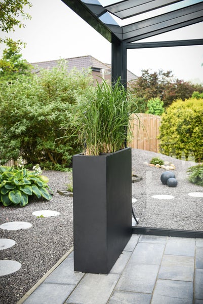 Hoher rechteckiger Pflanztrog in Schwarz mit Ziergras in einem modernen Garten mit Kies und Steinplatten.