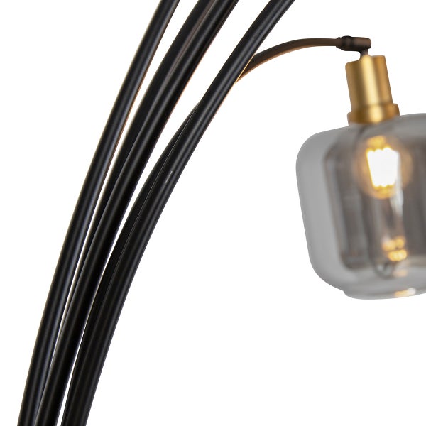 Nahaufnahme einer Stehleuchte mit schwarzen Metallarmen, goldener Fassung und rauchgrauem Glasschirm inklusive leuchtendem Filament-Leuchtmittel.