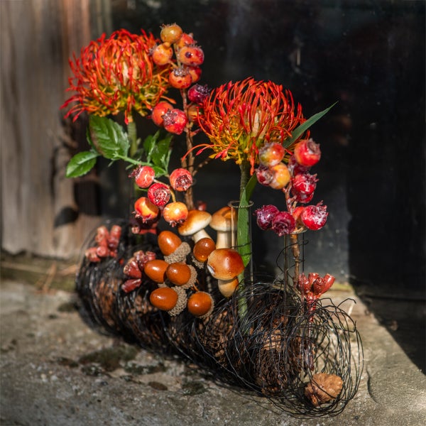 Herbstliches Gesteck mit Kunstblumen, Beeren, Pilzen und Eicheln in einem Drahtgeflecht mit Glasröhrchen.