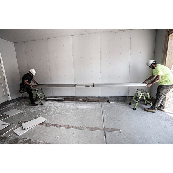 Zwei Handwerker mit Schutzhelmen auf einer Baustelle, die ein langes Aluminium Arbeitsgerüst auf zwei kleinen Stehleitern montieren.