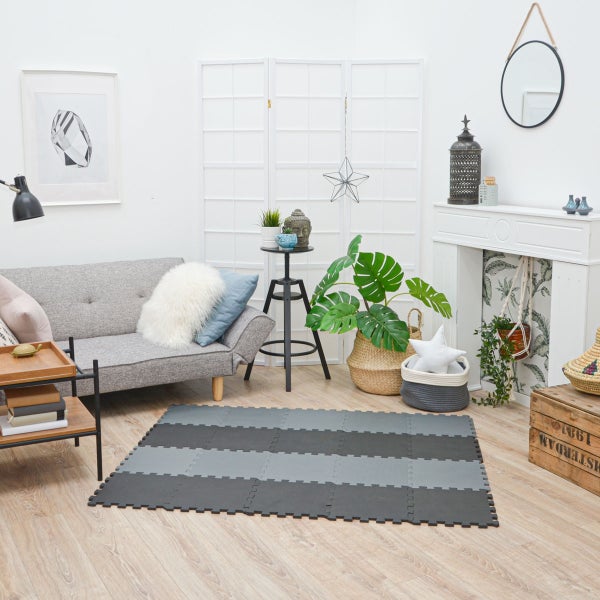 Puzzlematte aus Schaumstoff in Grau und Schwarz in einem hellen Wohnzimmer mit Sofa und Zimmerpflanzen auf Holzboden.