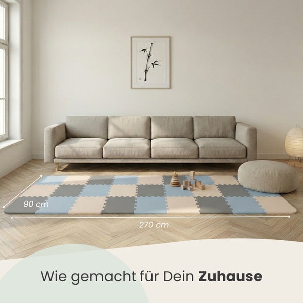Eine große Puzzlematte aus Schaumstoff in Blau, Grau und Beige auf einem Holzfußboden vor einem Sofa. Maße: 270 mal 90 Zentimeter.