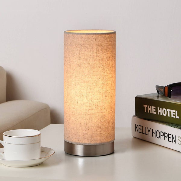 Zylindrische Tischleuchte mit beigem Stoffschirm und silbernem Metallsockel, leuchtend auf einem Tisch neben einer Tasse und Büchern.