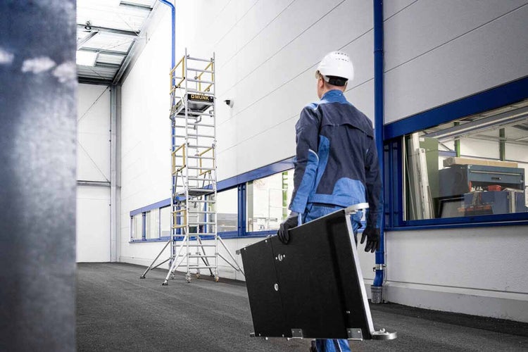 Ein Arbeiter trägt ein Plattformelement zu einem hohen fahrbaren Aluminium-Rollgerüst in einer hellen und organisierten Industriehalle.