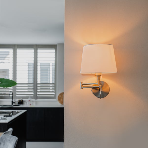 Eingeschaltete Wandleuchte mit weißem Stoffschirm und schwenkbarem Metallarm, montiert an einer hellen Wand.