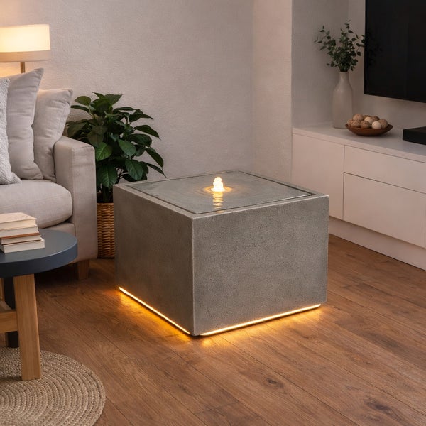 Würfelförmiger Zimmerbrunnen in Steinoptik mit LED-Beleuchtung am Boden und sprudelndem Wasser in einem modern gestalteten Wohnzimmer mit Holzboden.