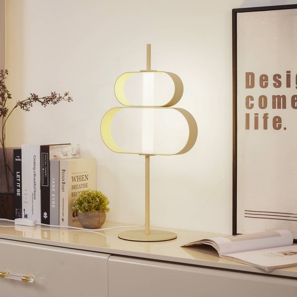 Moderne Tischleuchte aus Metall mit zwei geschwungenen Schlaufen als Lampenschirm auf einem Sideboard neben Büchern in einem hellen, organisierten Raum.