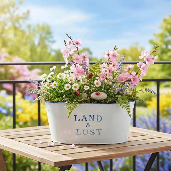 Weißer Metall-Pflanztopf mit Aufdruck Land und Lust, bepflanzt mit rosa Blüten, Gänseblümchen und Farnen auf einem Gartentisch aus Holz.
