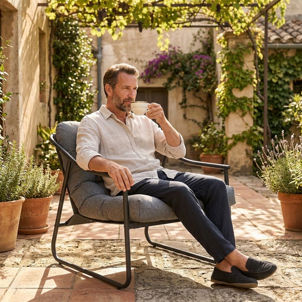 Mann sitzt in einem Gartenstuhl mit grauen Polstern auf einer sonnigen Terrasse mit vielen Pflanzen in Tontöpfen.