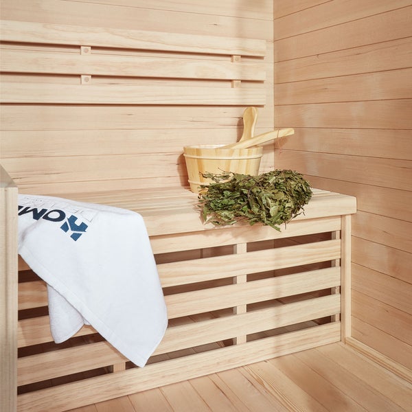 Holzsauna Innenraum mit Saunabank, Holzeimer, Kelle, Birkenreisig und einem Handtuch mit Hornbach Logo auf einer hellen Holzverkleidung.