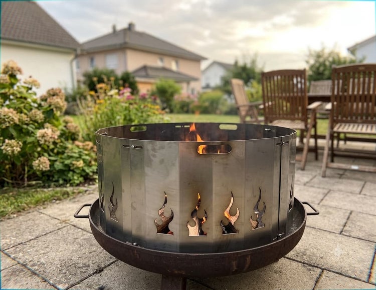 Feuerschale mit aufgesetztem Windschutz aus Metall mit flammenförmigen Aussparungen auf einer Terrasse im Garten.
