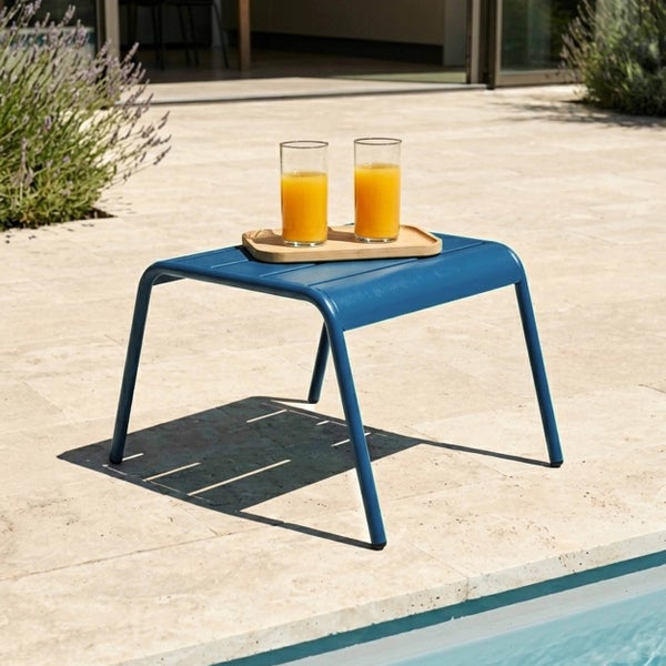 Niedriger Gartenhocker aus blauem Metall als Beistelltisch auf einer hellen Steinterrasse an einem Pool.