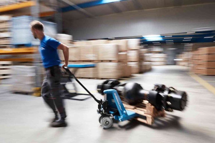 Mann zieht einen blauen Hanselifter Hubwagen mit einem schweren Maschinenteil auf einer Holzpalette in einem organisierten Lagerhaus.