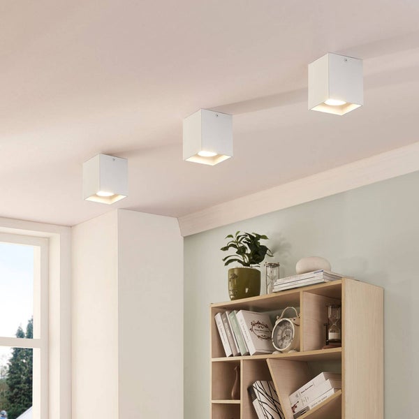 Drei weiße, würfelförmige Deckenleuchten an der Zimmerdecke über einem Regal