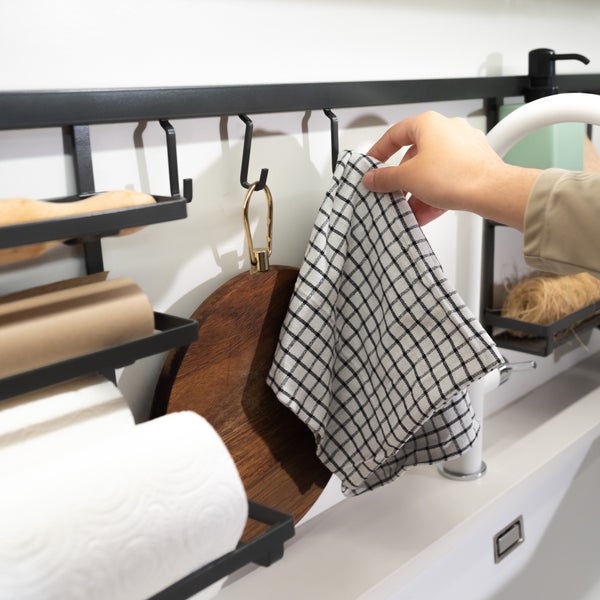Schwarze Küchenreling mit Papierrollenhalter, Schneidebrett und kariertem Geschirrtuch über einer Spüle.