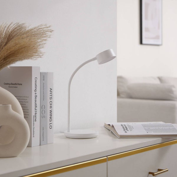 Moderne weiße LED-Schreibtischlampe mit biegsamem Hals auf einer weißen Ablage in einem hellen, aufgeräumten Wohnbereich neben Büchern.
