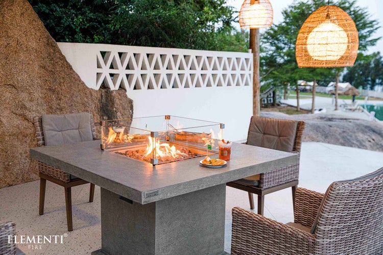 Quadratischer Gas-Feuertisch aus Faserbeton mit Glas-Windschutz, umgeben von Rattansesseln auf einer hellen, organisierten Terrasse.
